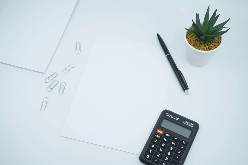 Calcolatrice, foglio bianco e penna nera appoggiati su una scrivania bianca moderna accanto ad una piantina grassa verde scuro. Gli strumenti per tenere d'occhio le spese e risparmiare sul caro bollette