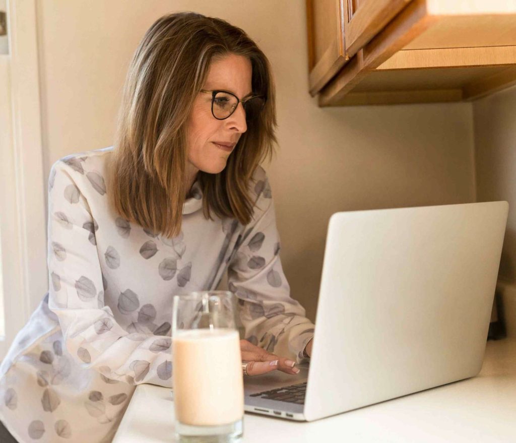 Donna con lunghi capelli castani e occhiali che vuole iniziare un'attività secondaria con il suo computer appoggiata al bancone della cucina