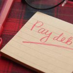 Primo piano di un blocchetto di post it a righe con scritto "Pay debt" uscire dai debiti, con accanto un pennarello rosso