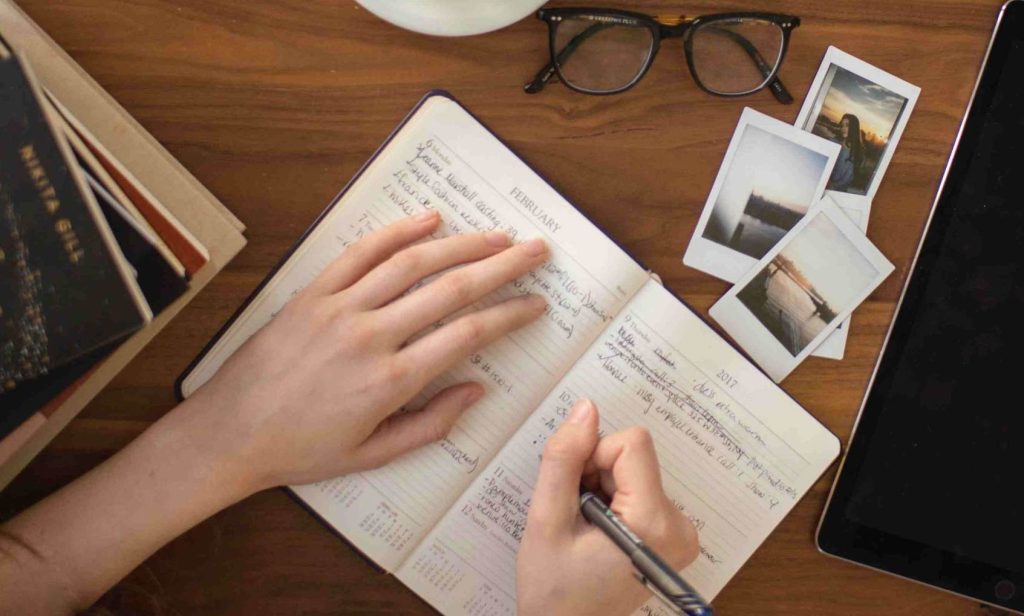Dettaglio di mani di giovane donna che sta effettuando registrazioni sul suo diario finanziario ad una scrivania di legno scuro con accanto alcune foto, occhiali, un tab e vecchi libri