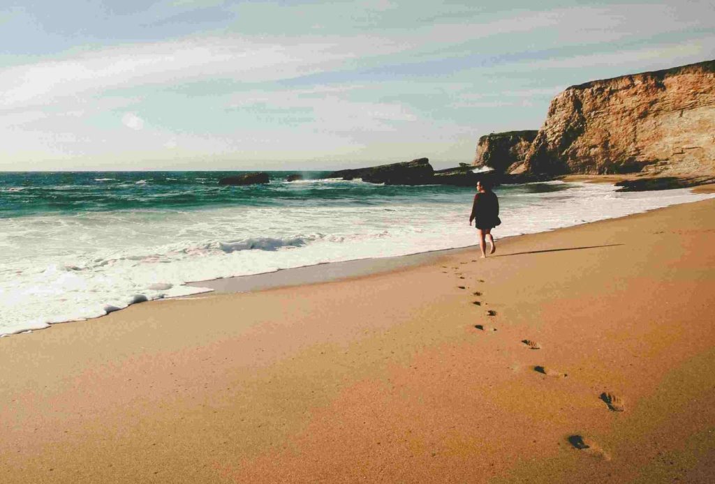 Donna che cammina incontro al mare su una spiaggia lasciando impronte nella sabbia dorata, sullo sfondo il cielo nei colori rosa dell'alba e una grande scogliera a picco. Per migliorare il rapporto con il denaro fai un passo alla volta