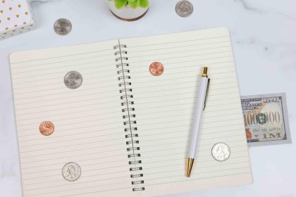Quaderno a righe a spirale con monetine, banconote e una penna bianco e oro su una superfice di marmo bianco. Annotare spese e risparmi è ottimo per capire perchè non riesci a risparmiare