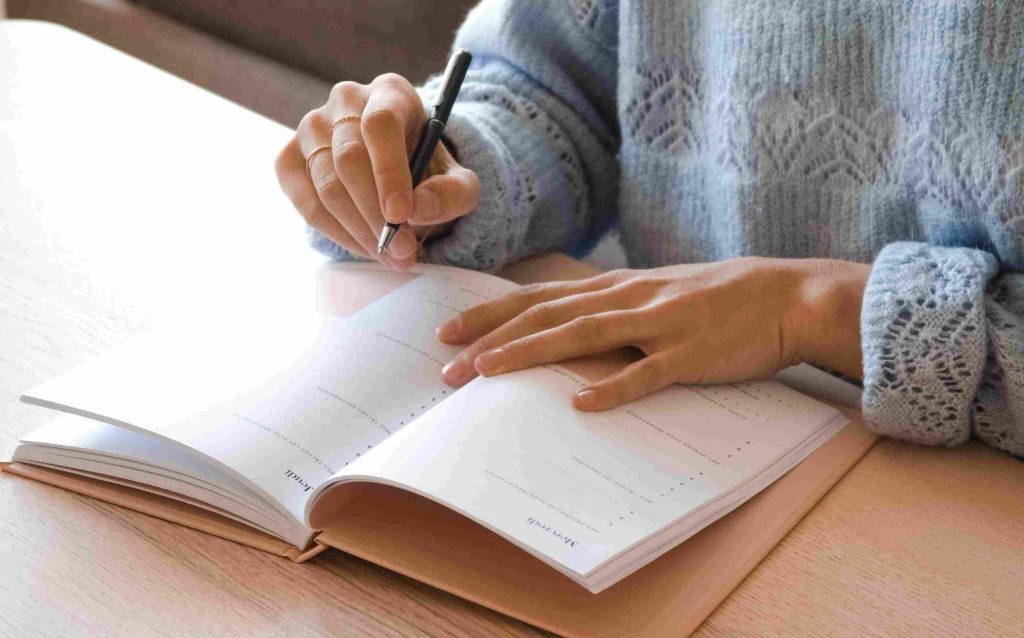 Giovane donna in maglione grigio traforato alla sua scrivania di legno chiaro che sta prendendo appunti per capire perché non riesce a risparmiare
