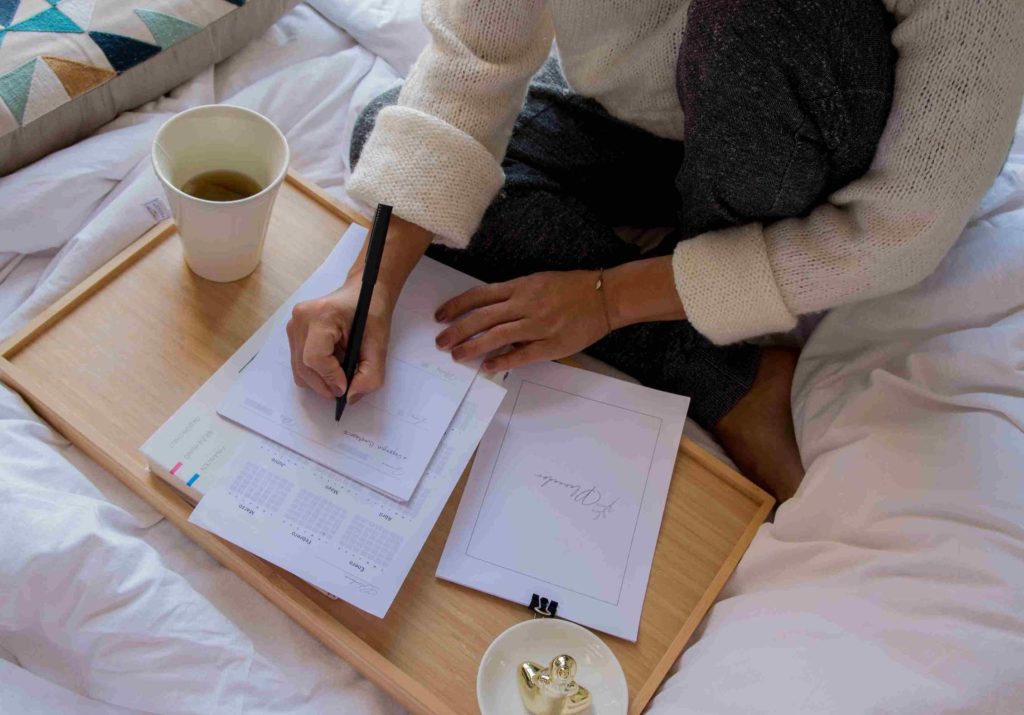 Donna in maglione chiaro e pantaloni scuri seduta sul suo letto che consulta i suoi documenti contabili con un buon te. Un ottimo modo per combattere la recessione