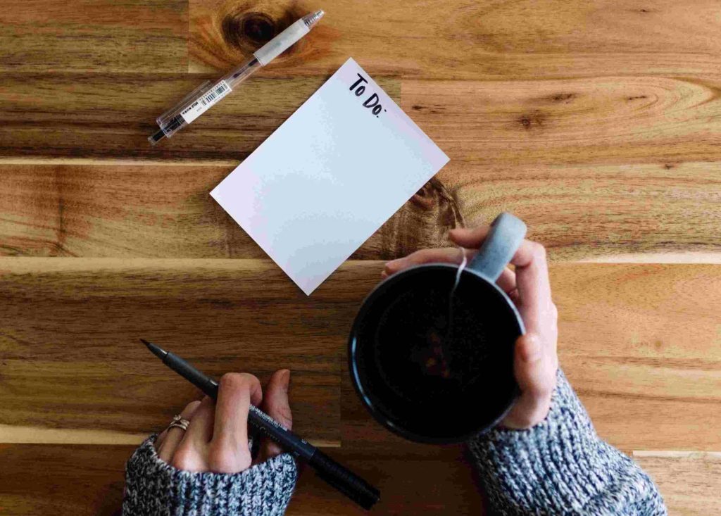 Carta penna e una buona tazza di caffè e compilare la tua to do list per risparmiare soldi velocemente come sta facendo la donna in maglione grigio alla sua scrivania di legno color miele nell'immagine