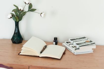 Immagine di una scrivania in legno chiaro con sopra un vaso nero con fiori bianchi, una boccetta di inchiostro vintage una pila di libri e un diario di lettura aperto in primo piano con una penna stilografica poggiata sopra